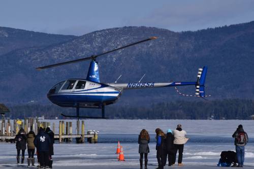 A blue and white helicopter hovers above a frozen lake, with people standing on the shore and a mountain backdrop.