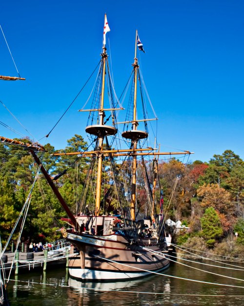 A tall ship with two masts docked by a wooded area under a clear blue sky.