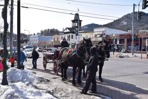 A horse-drawn carriage waits on a snowy street, with people walking and shops in the background. Mountains are visible.