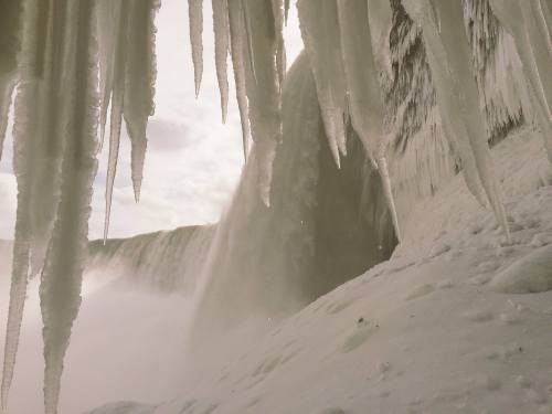Icicles hang from a rocky ledge, with a frozen waterfall cascading in the background under a cloudy sky.
