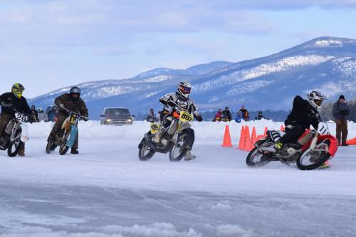 Motorcycles racing on ice with mountains in the background and spectators watching from a distance.