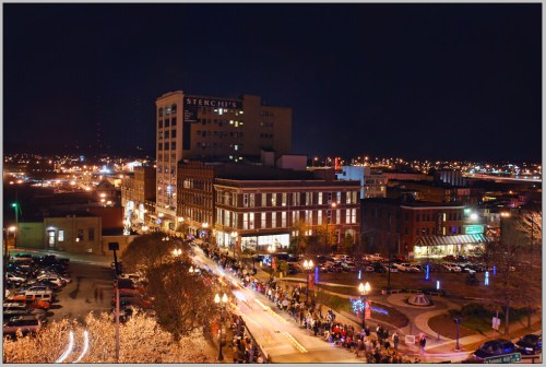 A bustling city street at night, lined with festive lights and crowds, with buildings and cars illuminated in the background.