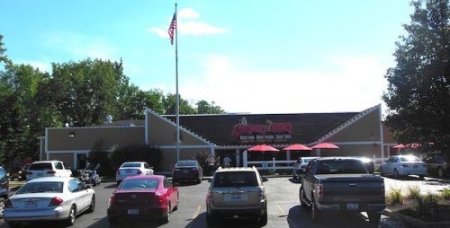Exterior view of a BBQ restaurant with a flag, parked cars, and outdoor seating under red umbrellas.