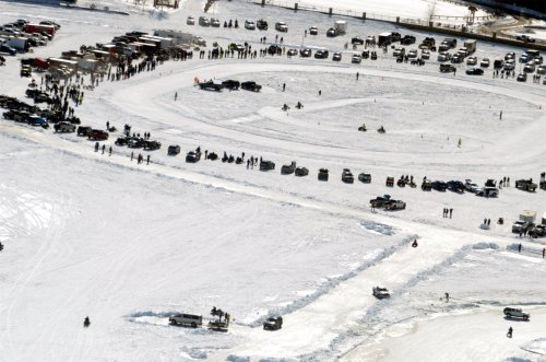 Aerial view of a snowy landscape with cars and people gathered around a circular track, likely for a winter event.
