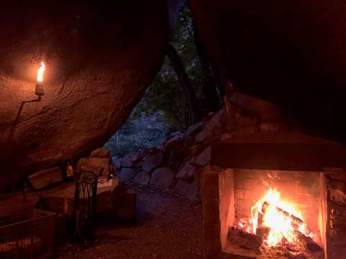 A cozy campsite with a fire burning in a stone fireplace, illuminated by a lantern and surrounded by large boulders.
