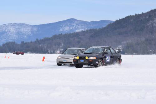 Two cars racing on a snowy landscape, with mountains in the background and orange cones marking the course.