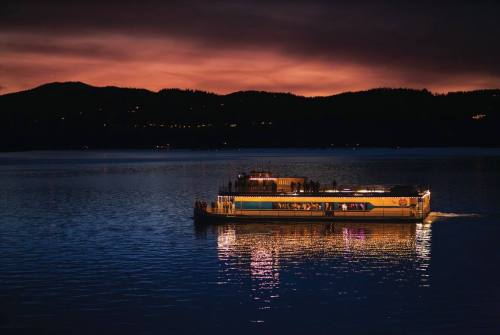 A boat illuminated at night glides across a calm lake, with mountains and a colorful sunset in the background.