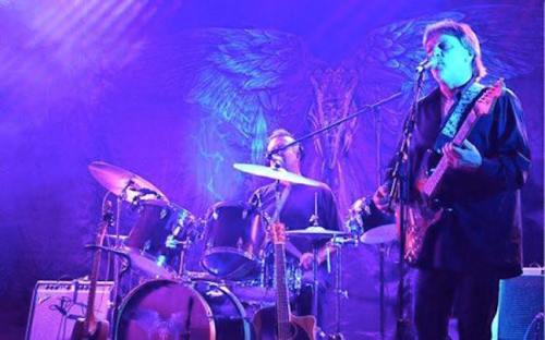 Two musicians perform on stage with drums and guitar, illuminated by purple lights and a backdrop featuring angelic wings.