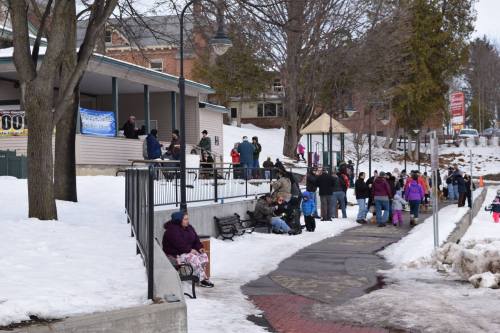 A snowy park scene with people gathered near a building, some sitting on benches and others standing in groups.