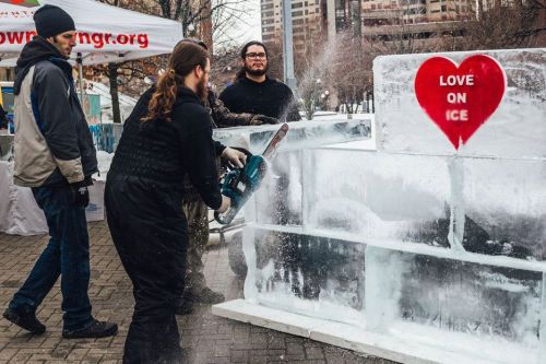 A person using a chainsaw to carve ice at an outdoor event, with a heart-shaped sign reading "Love on Ice."