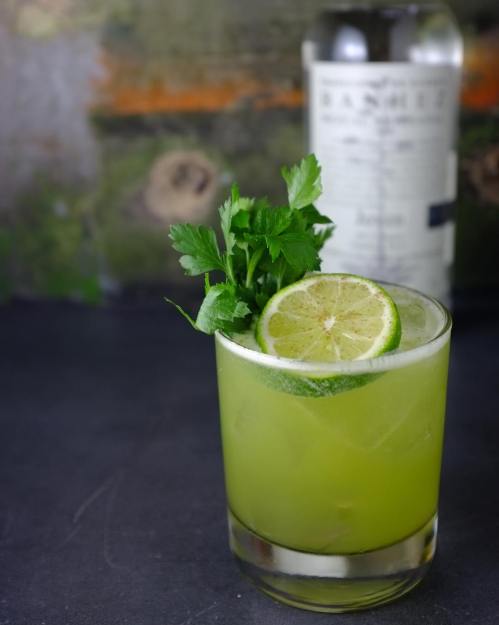 A refreshing green cocktail garnished with lime and cilantro, with a bottle of spirits in the background.