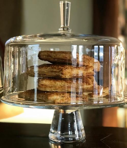 A glass cake dome displaying a stack of golden, flaky pastries on a pedestal.