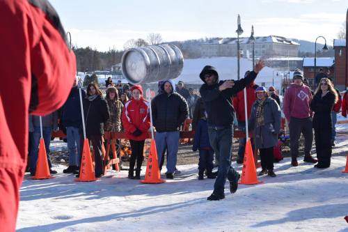 A person in a black hoodie throws a keg while a crowd watches, surrounded by snow and orange cones.