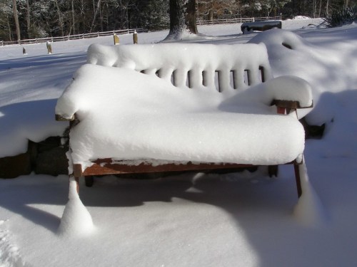 A wooden bench covered in a thick layer of snow, surrounded by a winter landscape.