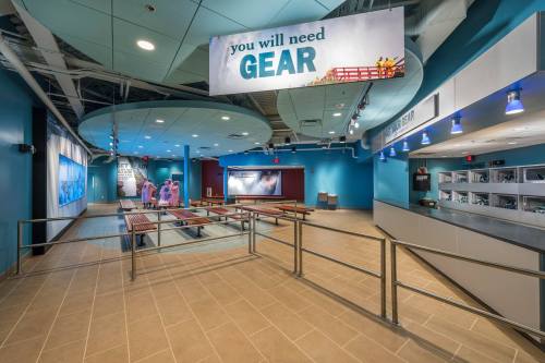 Interior view of a modern facility with blue walls, a sign reading "you will need GEAR," and various display areas.