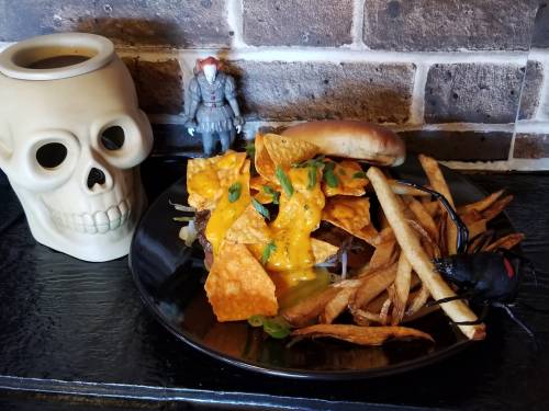 A plate with a loaded burger topped with nachos and cheese, fries, a skull mug, and a toy clown figure.