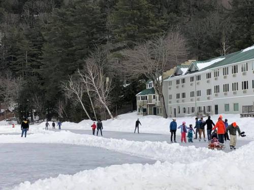 People ice skating on a frozen lake, with snow-covered banks and a building in the background. Trees line the area.