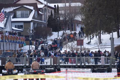 A large crowd gathers on a snowy street near a lake, with people on snowmobiles and buildings in the background.