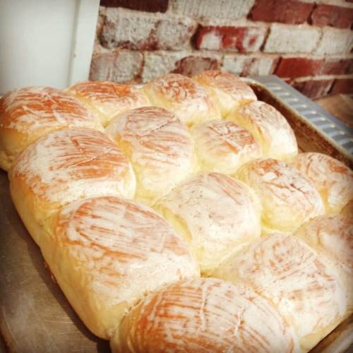 Freshly baked golden-brown dinner rolls arranged in a tray, with a rustic brick wall in the background.