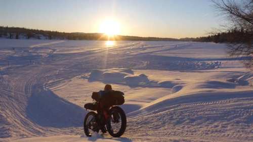A fat bike rests on a snowy landscape at sunset, with tracks in the snow and a serene lake in the background.