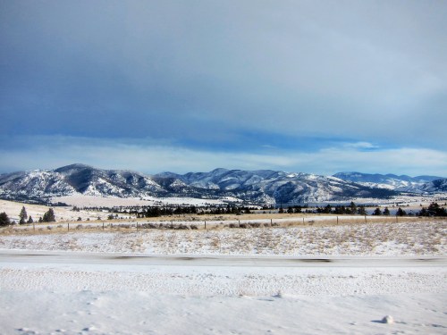 Snow-covered mountains under a cloudy sky, with a foreground of white fields and sparse trees.