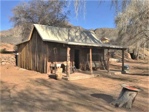A rustic wooden cabin with a metal roof, surrounded by dry landscape and mountains in the background.
