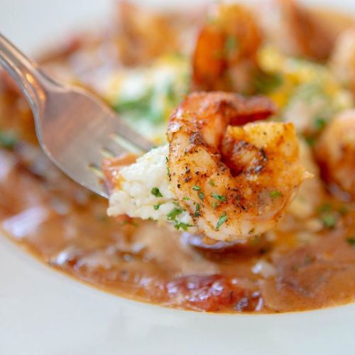 A fork holding a shrimp above a plate of shrimp and grits, garnished with herbs and a rich sauce.