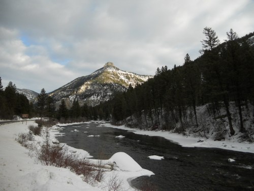 A snowy riverbank with a flowing river, surrounded by pine trees and mountains under a cloudy sky.