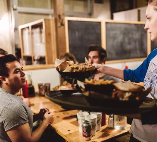 A server delivers food on a tray to a diner at a casual restaurant with others seated at the table.