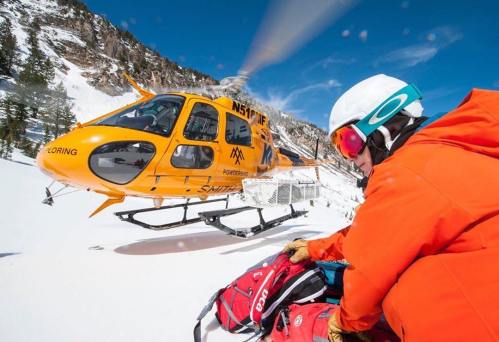 A skier in an orange jacket prepares gear near a bright orange helicopter on a snowy mountain landscape.