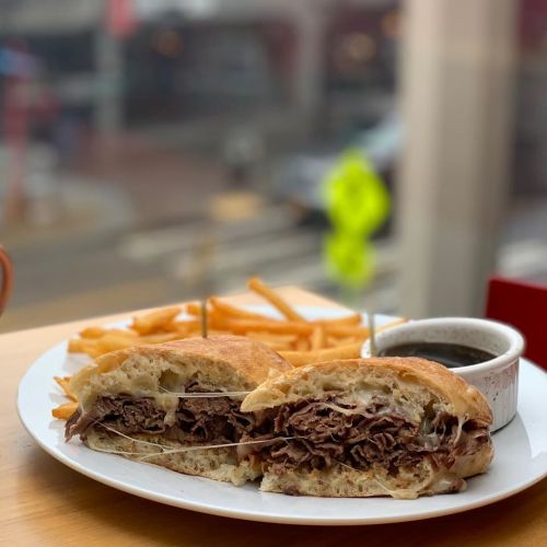 A close-up of a sliced sandwich filled with beef and melted cheese, served with fries and a small bowl of dipping sauce.