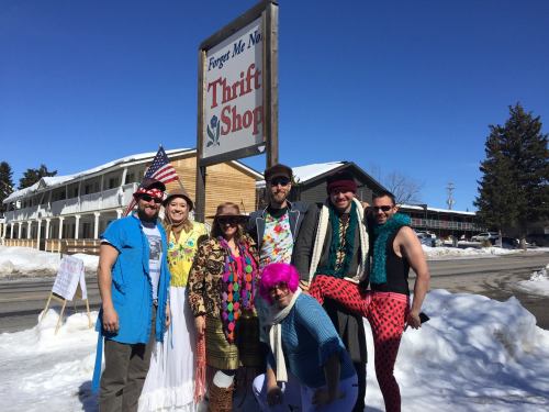 A group of people in colorful outfits poses outside a thrift shop sign, with snow and buildings in the background.