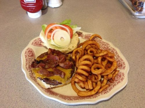 A plate with a cheeseburger topped with bacon, lettuce, tomato, onion, and curly fries on the side.