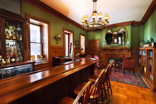 A cozy bar area with wooden furniture, green walls, a chandelier, and a fireplace, featuring shelves of drinks and decor.