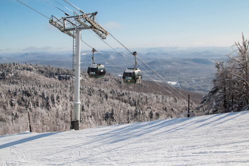 Two ski gondolas suspended above a snowy landscape, with trees and mountains in the background under a clear blue sky.