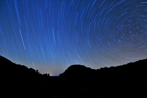 A night sky with star trails forming circular patterns over a dark landscape, creating a serene celestial scene.