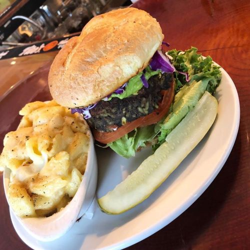 A hearty burger with lettuce, tomato, and purple cabbage, served with creamy macaroni and cheese and a pickle slice.