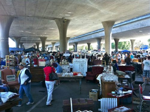 A bustling market scene under a highway, with vendors and shoppers among various stalls and goods.