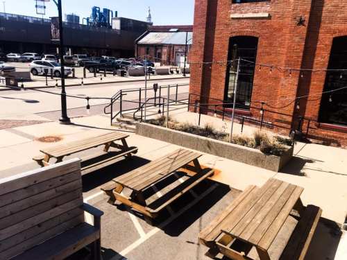 Outdoor seating area with wooden picnic tables and a landscaped planter, set against a brick building and urban backdrop.