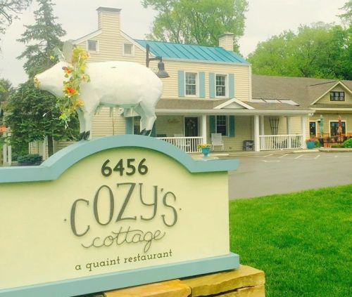 Sign for Cozy's Cottage, a quaint restaurant, featuring a white pig statue and a charming building in the background.