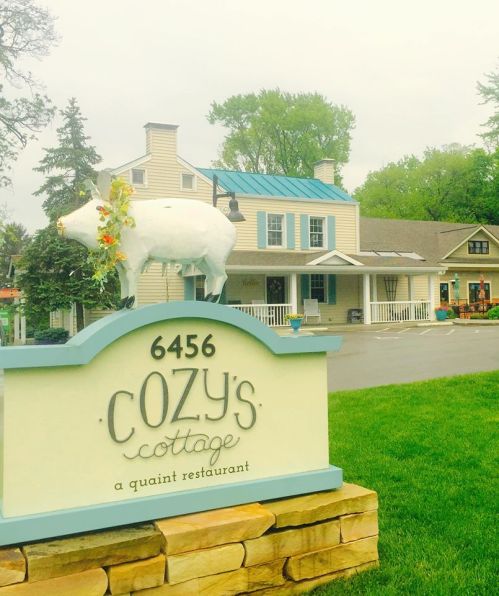 Sign for Cozy's Cottage, a quaint restaurant, featuring a decorative pig and a charming building in the background.