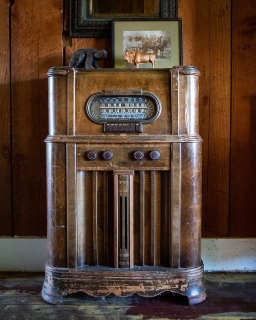 A vintage wooden radio with knobs, displayed on a wooden floor against a rustic wall, topped with a photo and a small bear figurine.