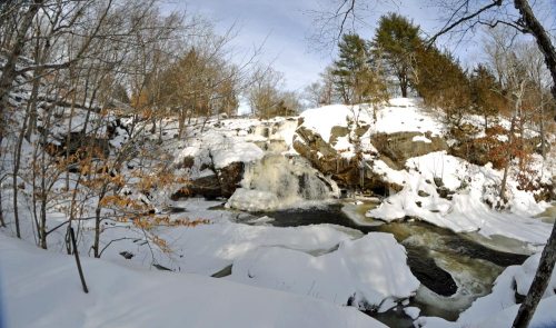 A snowy landscape featuring a waterfall cascading over rocks, surrounded by trees and a serene winter atmosphere.