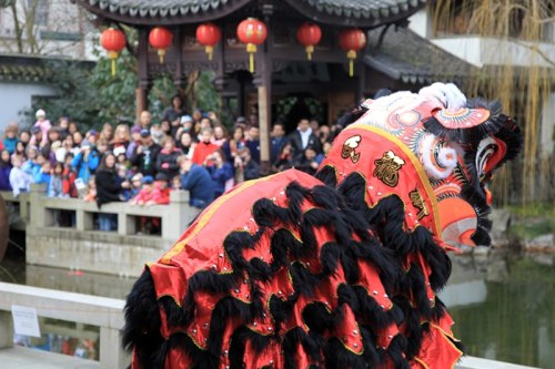 A lion dance performance in front of a large crowd near a pond, with traditional decorations and red lanterns.