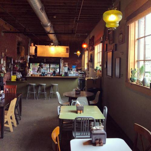 Cozy café interior with wooden beams, colorful lamps, and tables set for customers. Bar area in the background.