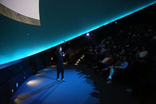 A speaker gestures on stage in a dimly lit auditorium, with an audience seated in the background.