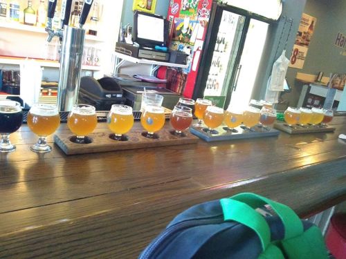 A wooden flight board with various craft beer samples lined up on a bar, with a green backpack in the foreground.