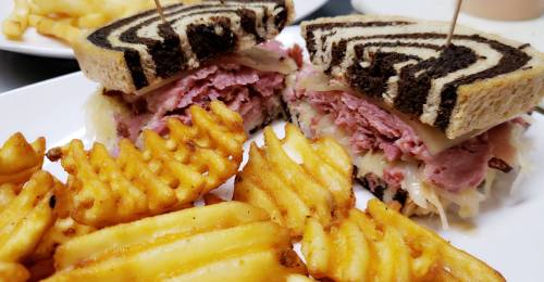 A close-up of a sliced sandwich with corned beef and sauerkraut on marbled rye, served with crinkle-cut fries.