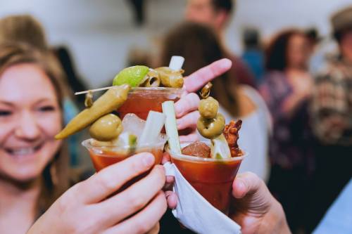A group cheers with colorful cocktails garnished with olives, celery, and lime in a lively social setting.