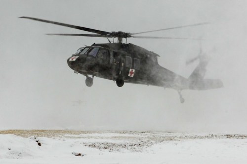 A military helicopter flying in snowy, foggy conditions, with a red cross symbol on its side.